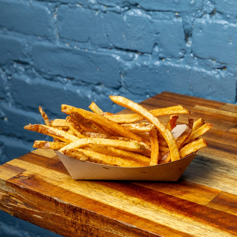 Golden-brown french fries in a paper tray on a wooden table, with a blue brick wall background.