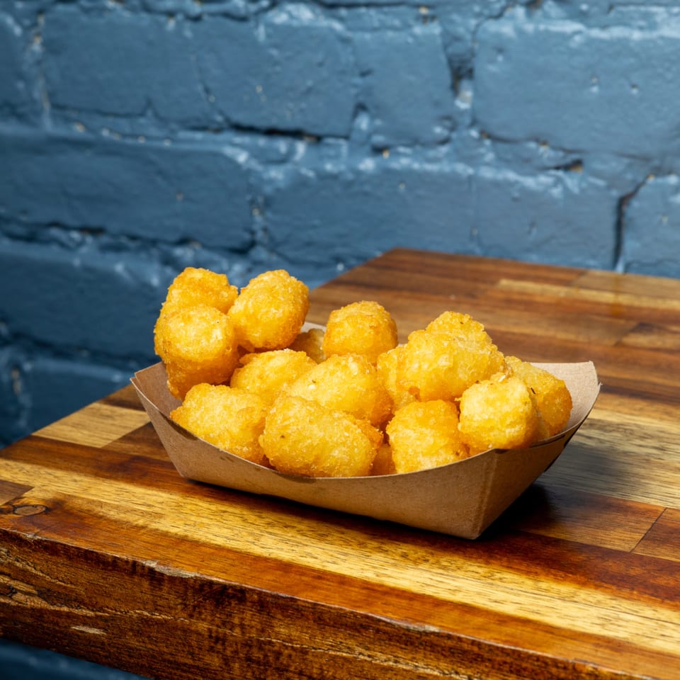 Golden brown tater tots in a paper tray on a wooden table, with a blue brick wall background.