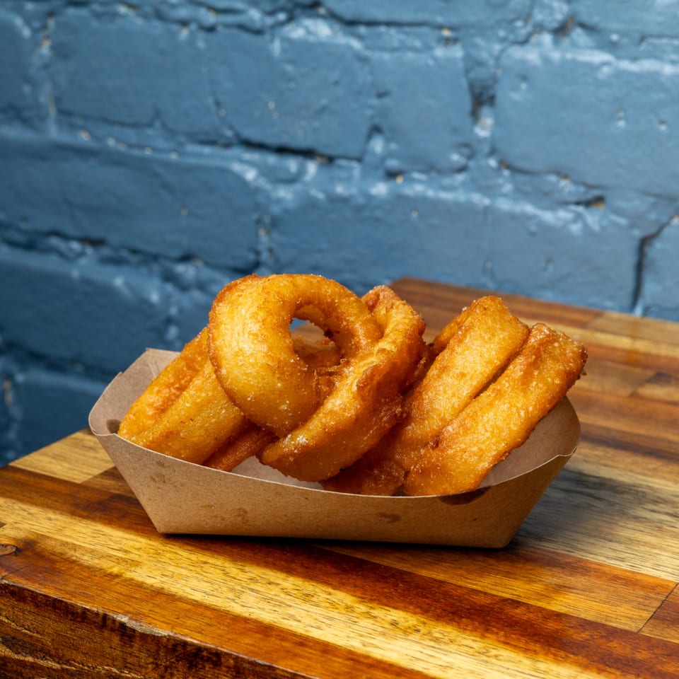 Golden-brown onion rings and fries in a paper tray on a wooden surface with a blue brick wall.