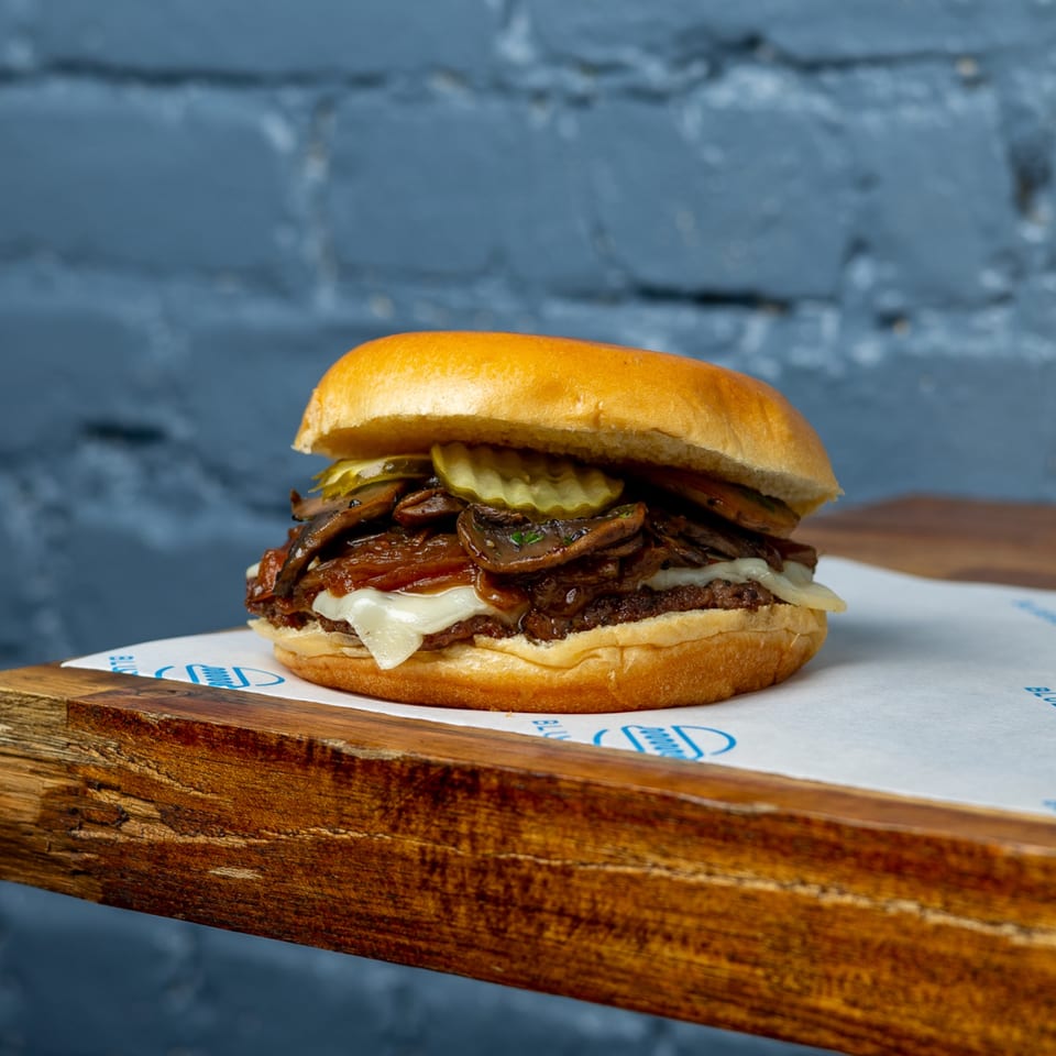 Blue Road Burger with mushrooms, caramelized onions, pickles, and melted cheese on a wooden board.
